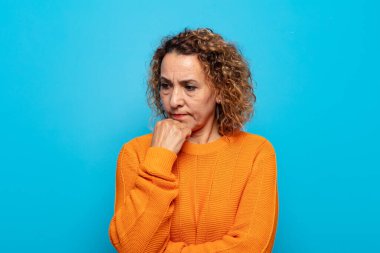 Orta yaşlı kadın ciddi, düşünceli ve endişeli, elleri çenesine bastırılmış bir şekilde yan bakıyor.