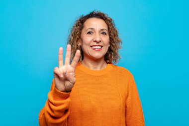 Orta yaşlı bir kadın gülümsüyor ve arkadaş canlısı görünüyor. Üç ya da üç numarayı gösteriyor. Elleri önde, geri sayıyor.