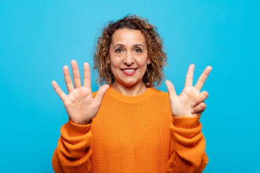 Orta yaşlı bir kadın gülümsüyor ve arkadaş canlısı görünüyor, 8 ya da 8 numarayı gösteriyor, elleri önde, geriye doğru sayıyor.