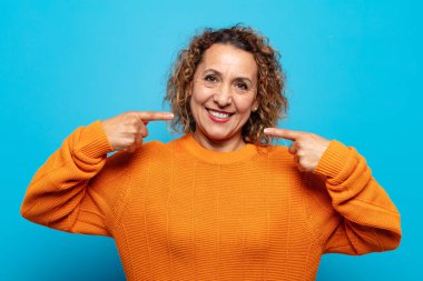 Orta yaşlı kadın kendine güvenerek gülümsüyor. Geniş bir gülümseme, pozitif, rahat, tatmin olmuş bir tavır.