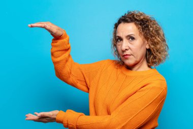 Orta yaşlı bir kadın bir objeyi iki eliyle fotokopi alanında tutuyor, bir objeyi gösteriyor, sunuyor ya da reklamını yapıyor.