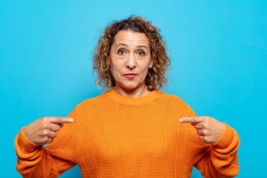 Orta yaşlı kadın gururlu, pozitif ve rahat görünüyor. İki eliyle göğsünü gösteriyor.