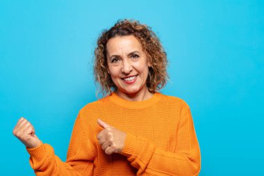 Orta yaşlı kadın neşeli bir şekilde gülümsüyor ve yan taraftaki fotokopi alanını işaret ediyor, mutlu ve tatmin olmuş hissediyor.