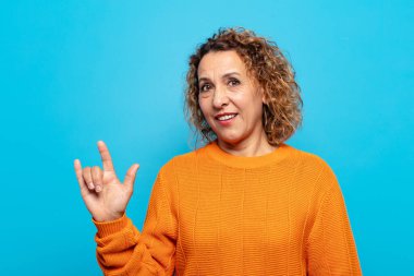 Orta yaşlı bir kadın mutlu, eğlenceli, kendinden emin, pozitif ve asi hissediyor, elle kaya veya heavy metal işareti yapıyor.