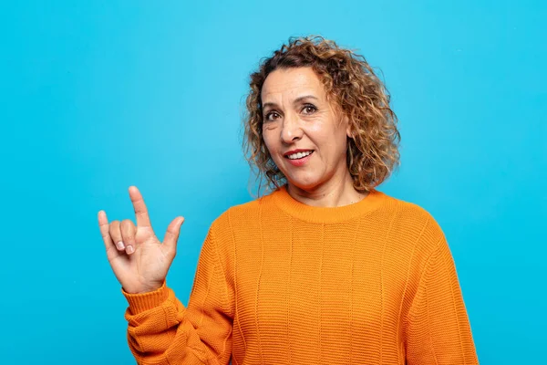 Orta yaşlı bir kadın mutlu, eğlenceli, kendinden emin, pozitif ve asi hissediyor, elle kaya veya heavy metal işareti yapıyor.