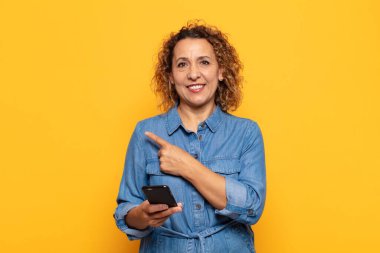 İspanyol orta yaşlı kadın neşeli bir şekilde gülümsüyor, mutlu hissediyor ve yan ve yukarı doğru işaret ediyor, fotokopi alanında nesneler gösteriyor.