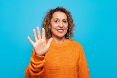 Orta yaşlı bir kadın gülümsüyor ve arkadaş canlısı görünüyor. 5 ya da 5 numarayı gösteriyor.