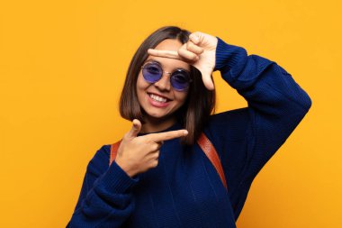 İspanyol kadın mutlu, arkadaş canlısı ve pozitif hissediyor, gülümsüyor ve elleriyle bir portre veya fotoğraf çerçevesi yapıyor.