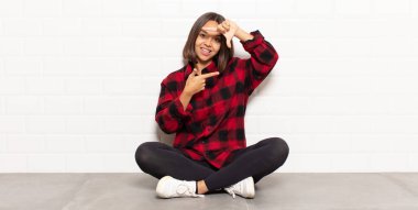 İspanyol kadın mutlu, arkadaş canlısı ve pozitif hissediyor, gülümsüyor ve elleriyle bir portre veya fotoğraf çerçevesi yapıyor.