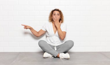 Orta yaşlı kadın kendini mutlu, şaşkın ve şaşırmış hissediyor. Ağzını eliyle kapatıyor ve yanal fotokopi alanını işaret ediyor.