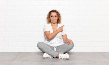 Orta yaşlı kadın neşeli bir şekilde gülümsüyor, mutlu hissediyor ve yan ve yukarı doğru işaret ediyor, fotokopi uzayındaki nesneleri gösteriyor.