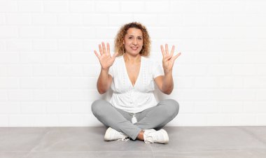 Orta yaşlı bir kadın gülümsüyor ve arkadaş canlısı görünüyor. 9 ya da 9 numarayı gösteriyor. Eli önde, geriye doğru sayıyor.