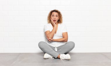 Orta yaşlı bir kadın şok ve inançsızlık içinde, elleri yanağında ve kolları çaprazlanmış, sersemlemiş ve şaşırmış hissediyor.