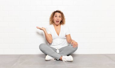 Orta yaşlı kadın şaşırmış ve şoka uğramış görünüyor, çenesi düşmüş ve eli açık bir nesneyi tutuyor.