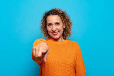 Orta yaşlı bir kadın kameraya bakıyor, kendinden emin, arkadaş canlısı bir gülümsemeyle, seni seçiyor.