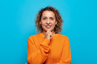 Orta yaşlı kadın mutlu görünüyor ve gülümsüyor. Elleri çenesinde, merak ediyor ya da soru soruyor. Seçenekleri karşılaştırıyor.