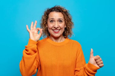 Orta yaşlı bir kadın mutlu, şaşkın, tatmin olmuş ve şaşırmış hissediyor, iyi görünüyor ve el kol hareketleri yapıyor, gülümsüyor.