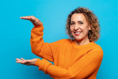 Orta yaşlı kadın gülümsüyor, mutlu, pozitif ve tatmin olmuş hissediyor, fotokopi alanında nesne veya konsept tutuyor veya gösteriyor