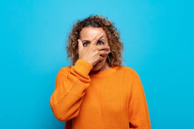 Orta yaşlı bir kadın şok olmuş, korkmuş ya da korkmuş görünüyor. Yüzü elle kaplanmış ve parmaklarının arasından gizlice bakıyor.