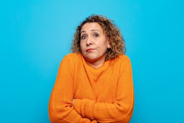 Orta yaşlı bir kadın omuz silkiyor, kafası karışık ve kararsız, kolları bağlı ve şaşkın görünüyor.