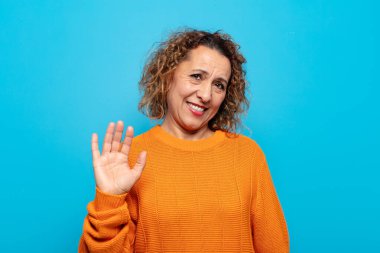 Orta yaşlı bir kadın mutlu ve neşeli bir şekilde gülümsüyor, el sallıyor, sizi selamlıyor ve selamlıyor, ya da veda ediyor.