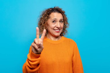 Orta yaşlı bir kadın gülümsüyor ve arkadaş canlısı görünüyor. İki ya da iki numarayı gösteriyor. Elleri önde, geriye doğru sayıyor.