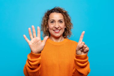 Orta yaşlı bir kadın gülümsüyor ve arkadaş canlısı görünüyor, altı ya da altıncı numarayı gösteriyor, eli önde, geriye doğru sayıyor.