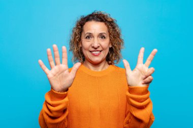 Orta yaşlı bir kadın gülümsüyor ve arkadaş canlısı görünüyor, 8 ya da 8 numarayı gösteriyor, elleri önde, geriye doğru sayıyor.