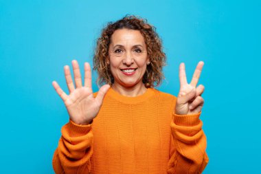 Orta yaşlı bir kadın gülümsüyor ve dost canlısı görünüyor, 7 ya da 7 numarayı gösteriyor, eli önde, geriye doğru sayıyor.