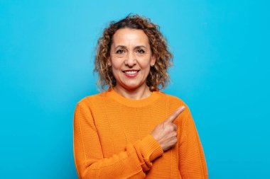 Orta yaşlı kadın neşeli bir şekilde gülümsüyor, mutlu hissediyor ve yan ve yukarı doğru işaret ediyor, fotokopi uzayındaki nesneleri gösteriyor.