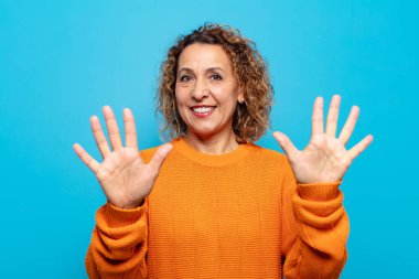 Orta yaşlı bir kadın gülümsüyor ve arkadaş canlısı görünüyor, onuncu veya onuncu numarayı gösteriyor, eli önde, geriye doğru sayıyor.