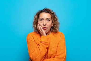 Orta yaşlı bir kadın şok ve inançsızlık içinde, elleri yanağında ve kolları çaprazlanmış, sersemlemiş ve şaşırmış hissediyor.