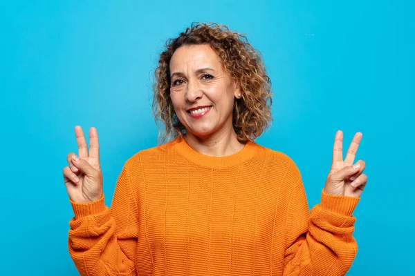 Orta yaşlı kadın gülümsüyor ve mutlu görünüyor, arkadaş canlısı ve memnun, zafer ya da barış işareti yapıyor iki eliyle