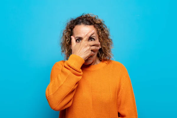 Orta yaşlı bir kadın şok olmuş, korkmuş ya da korkmuş görünüyor. Yüzü elle kaplanmış ve parmaklarının arasından gizlice bakıyor.