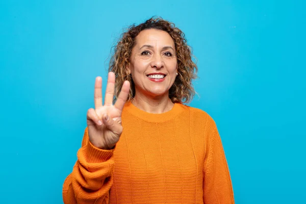 Orta yaşlı bir kadın gülümsüyor ve arkadaş canlısı görünüyor. Üç ya da üç numarayı gösteriyor. Elleri önde, geri sayıyor.