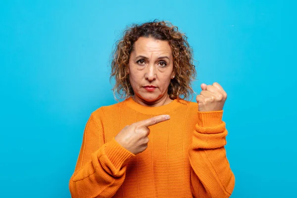 Orta yaşlı bir kadın sabırsız ve kızgın görünüyor, saati gösteriyor, dakiklik istiyor, zamanında gelmek istiyor.