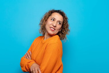 Orta yaşlı bir kadın neşeyle gülümsüyor, mutlu, tatmin olmuş ve rahatlamış hissediyor, kollarını kavuşturup yan tarafa bakıyor.