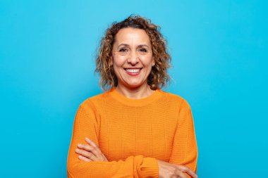 Orta yaşlı bir kadın mutlu, gururlu ve tatmin olmuş görünüyor. Kollarını kavuşturup gülümsüyor.