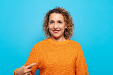 Orta yaşlı kadın gururlu, kendinden emin ve mutlu görünüyor, gülümsüyor ve kendini işaret ediyor ya da bir numara işareti yapıyor.