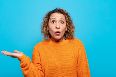 Orta yaşlı kadın şaşırmış ve şoka uğramış görünüyor, çenesi düşmüş ve eli açık bir nesneyi tutuyor.