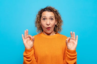 Orta yaşlı kadın şok olmuş, şaşırmış ve şaşırmış hissediyor. Onaylıyor. İki eliyle de onay işareti yapıyor.