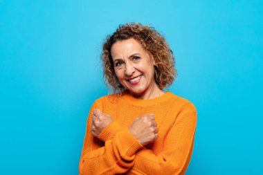 Orta yaşlı bir kadın neşeli bir şekilde gülümsüyor ve yumruklarını sıkmış, kollarını kavuşturmuş, mutlu ve pozitif hissediyor.