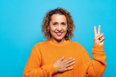 Orta yaşlı kadın mutlu, kendinden emin ve güvenilir görünüyor, gülümsüyor ve zafer işareti gösteriyor, olumlu bir tavırla.