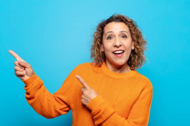 Orta yaşlı bir kadın mutlu ve şaşırmış hissediyor. Şok olmuş bir ifadeyle gülümsüyor ve bir tarafı işaret ediyor.