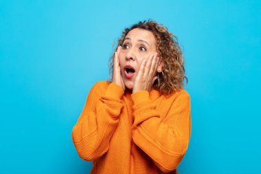 Orta yaşlı bir kadın, mutlu, heyecanlı ve şaşırmış hissediyor, iki eli yüzü yana bakıyor.