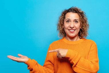 Orta yaşlı kadın neşeyle gülümsüyor ve yan taraftaki fotokopi alanına işaret ediyor, bir nesneyi gösteriyor veya reklamını yapıyor.