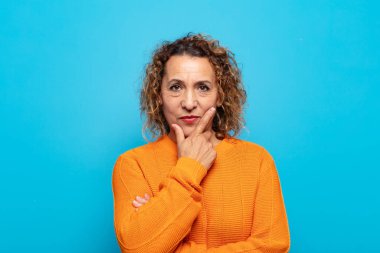 Orta yaşlı bir kadın ciddi, düşünceli ve güvensiz görünüyor, bir kolu çapraz ve çenesi üzerinde, seçenekleri tartıyor.