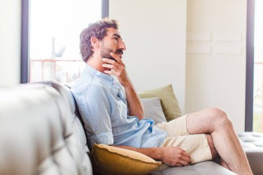 Genç sakallı adam evdeki kanepede oturuyor.