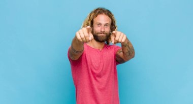 Sarışın adam mutlu ve kendinden emin, kamerayı iki eliyle gösteriyor ve gülüyor, seni seçiyor.