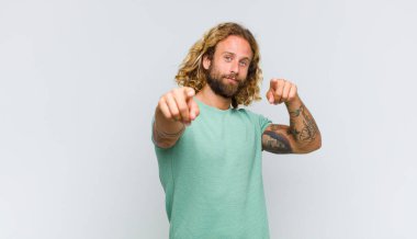 Sarışın adam mutlu ve kendinden emin, kamerayı iki eliyle gösteriyor ve gülüyor, seni seçiyor.